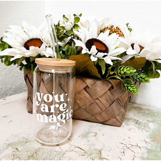 A glass can with a white vinyl design that says "you are magic". The glass can has a bamboo lid and a glass straw. In the background is a brown woven basket with white daisys and greenery.
